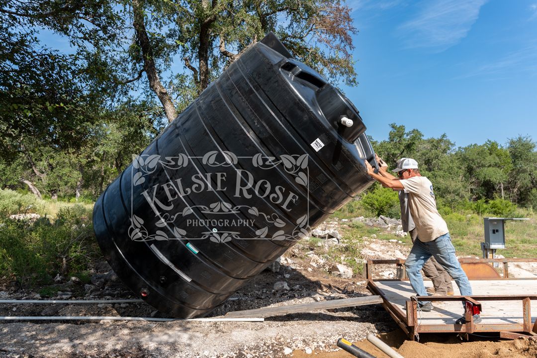 Man unloading a large black water storage tank from a trailer outdoors on a sunny day.