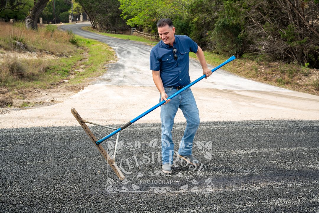 Smiling man in blue shirt raking gravel on a newly paved rural road.