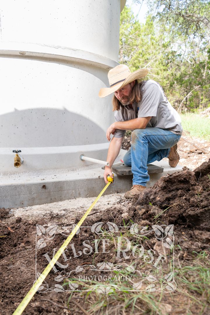 Man in cowboy hat using tape measure near base of large concrete water storage tank outdoors.