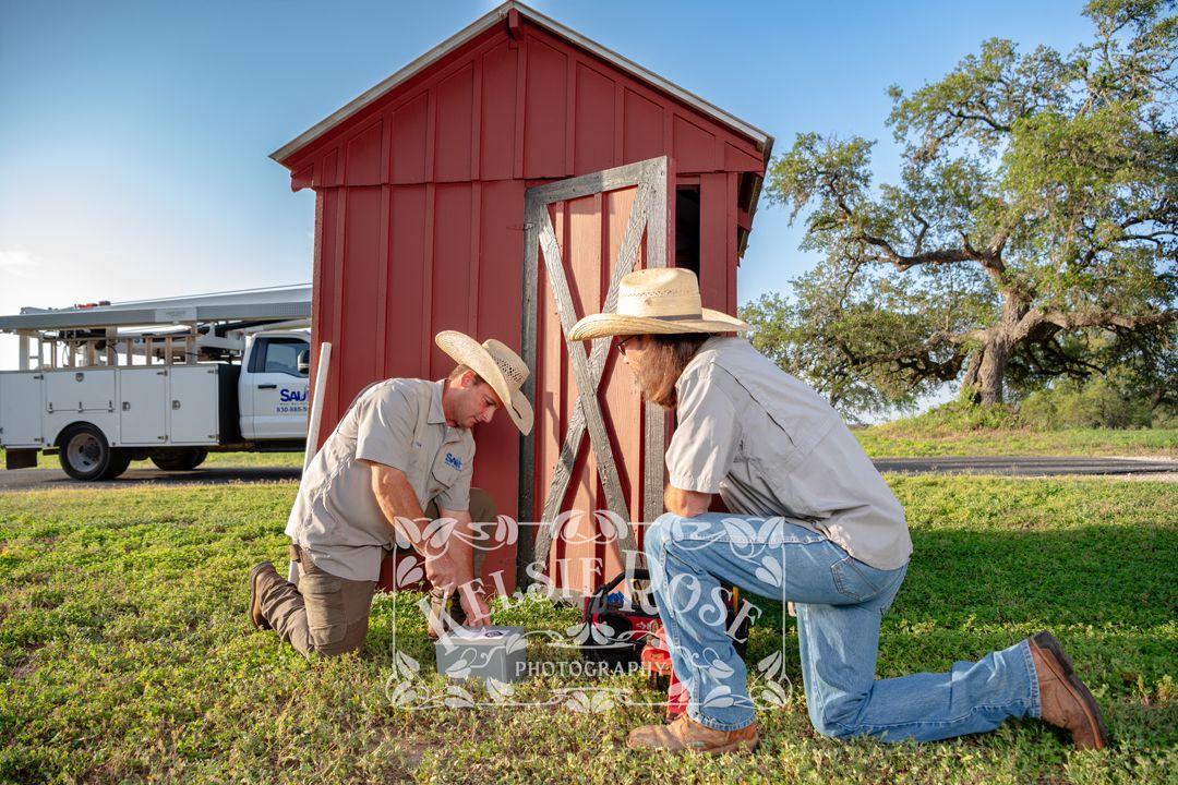 Two workers in cowboy hats kneeling on grass near a red well building, performing maintenance work on a sunny day.
