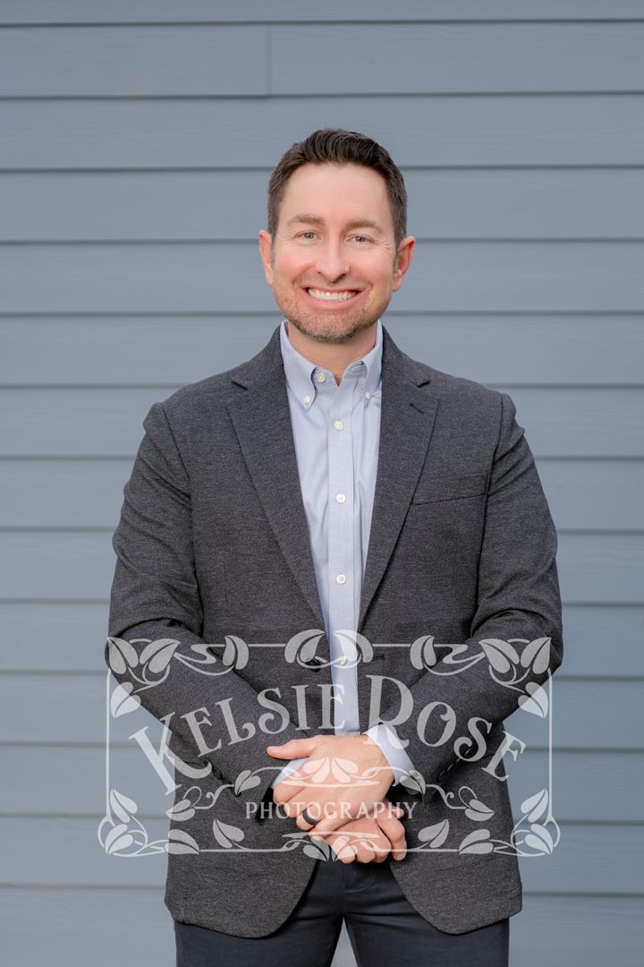 Smiling man in gray blazer and light blue shirt smiling with clasped hands against gray siding wall, professional portrait photography.