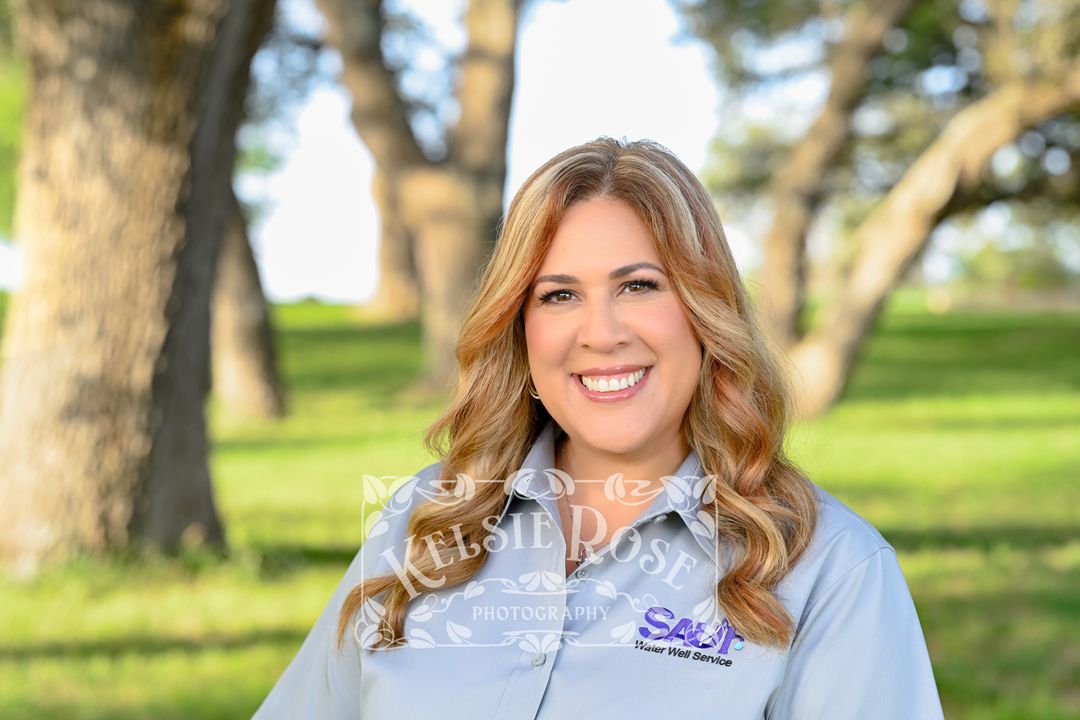 Headshot of smiling woman with brunette hair wearing water well company uniform shirt outdoors.