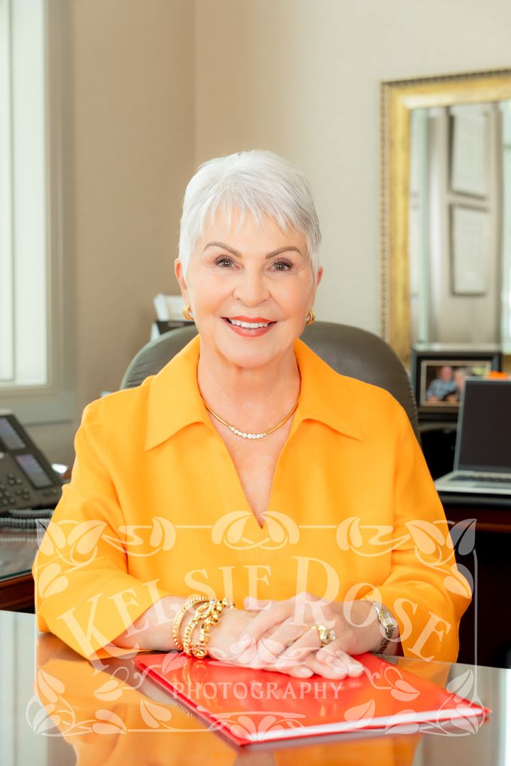 Elegant senior woman in orange blazer sitting confidently at office desk with hands folded.