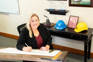 Blonde woman in black blazer sitting at drafting board with construction hard hats in office.