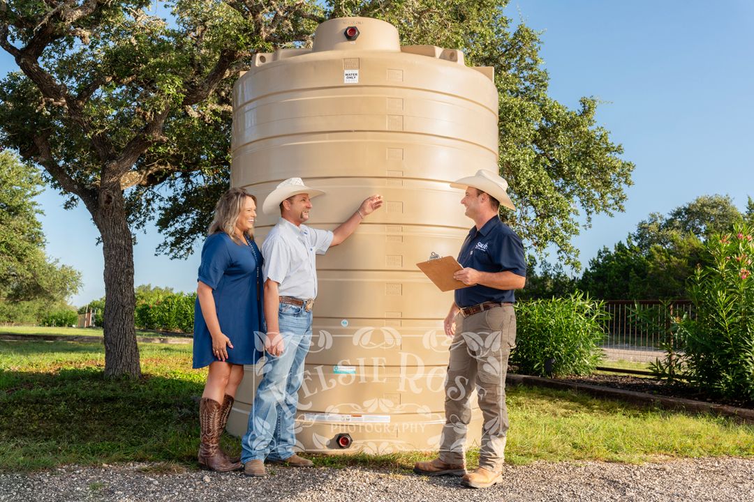 A couple of customers consult with a clipboard-holding business owner beside a large tan water storage tank outdoors.