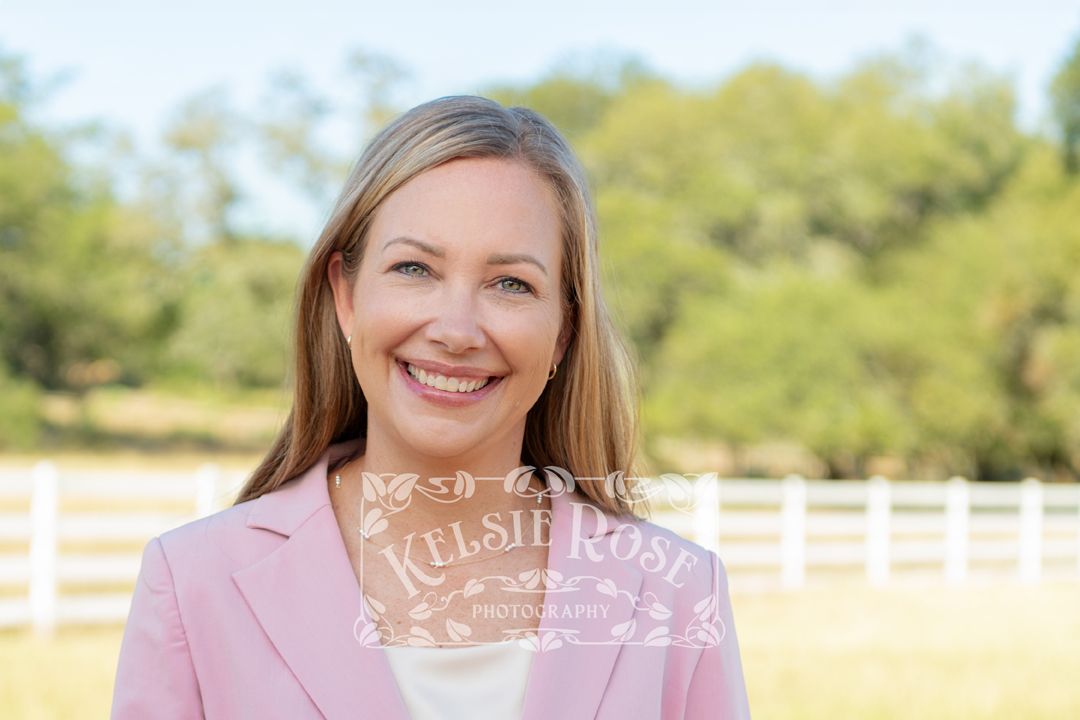 Headshot of smiling blonde woman in pink blazer poses outdoors near white fence with field in background.