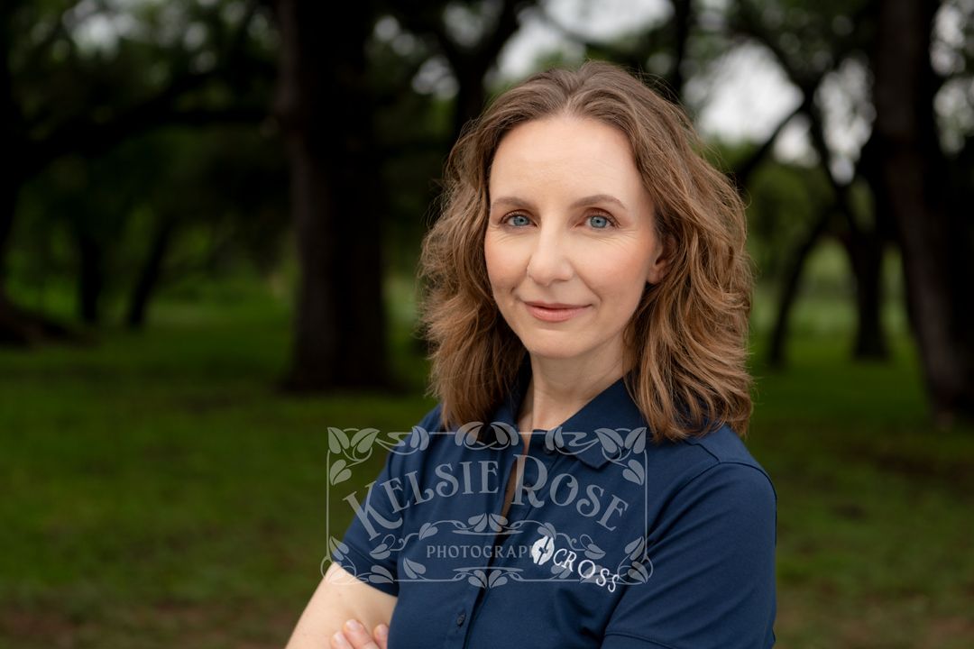 Headshot of woman with wavy brown hair poses outdoors in navy blue shirt, oak trees in background.