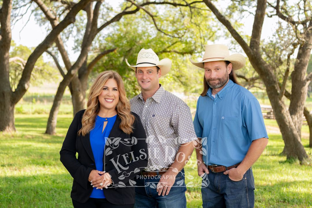 Three people pose outdoors among trees, two men in cowboy hats flank a smiling woman in a blue top in a professional portrait.