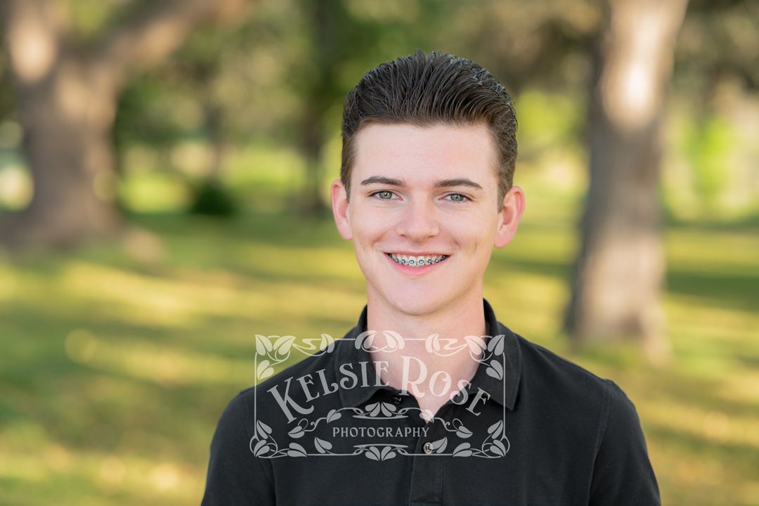 Smiling teenage boy with braces wearing a black shirt in a green outdoor setting.
