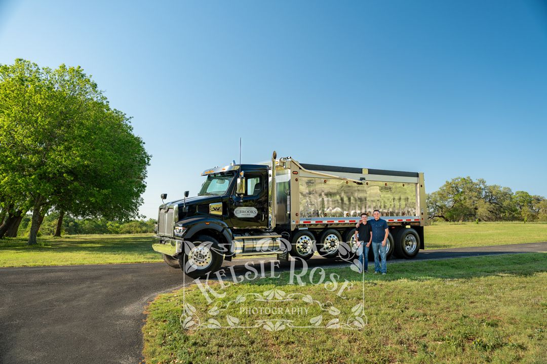Man standing beside a large black and chrome dump truck in a green ranch setting on a sunny day.