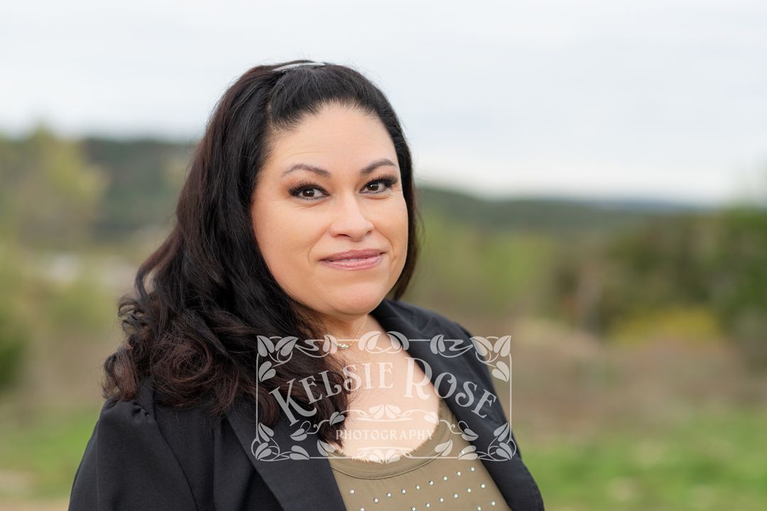 Headshot of woman with dark wavy hair smiling outdoors wearing black blazer and olive green top.