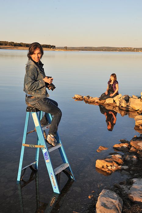 Female photographer sitting on blue ladder in the water holding camera near lake with subject posing on rocks during golden hour.