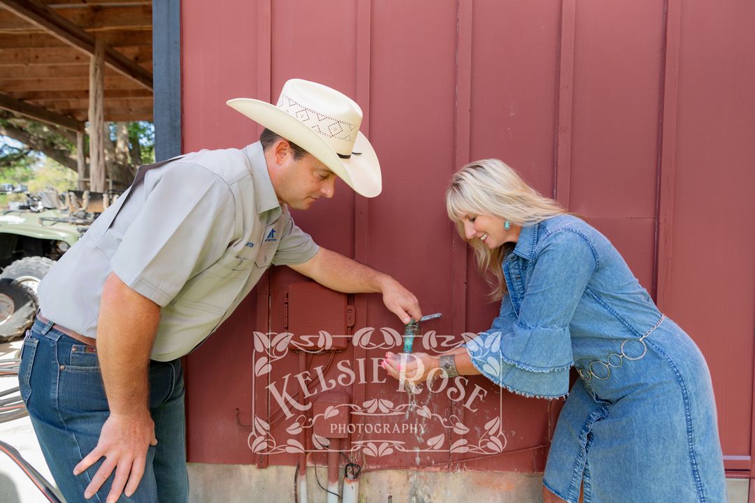 Man in cowboy hat turning outdoor faucet while customer washes her hands under running water outdoors near a red barn.