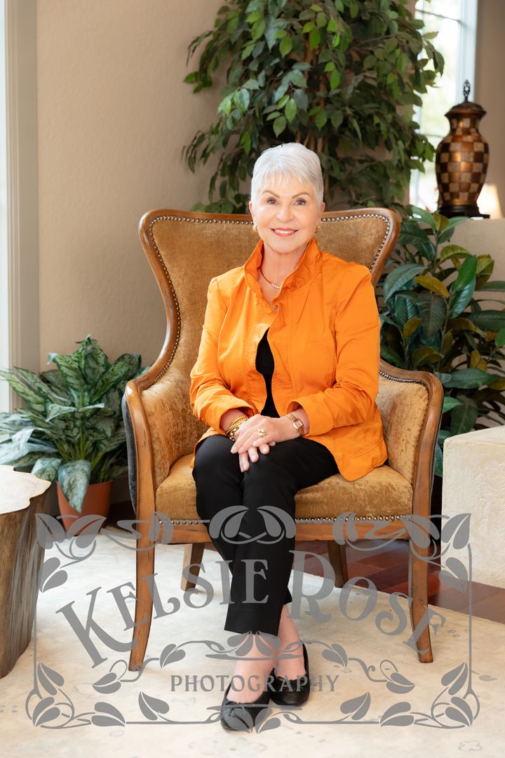 Elegant older woman in orange blazer sitting in gold wingback chair surrounded by lush green plants, professional portrait photo.