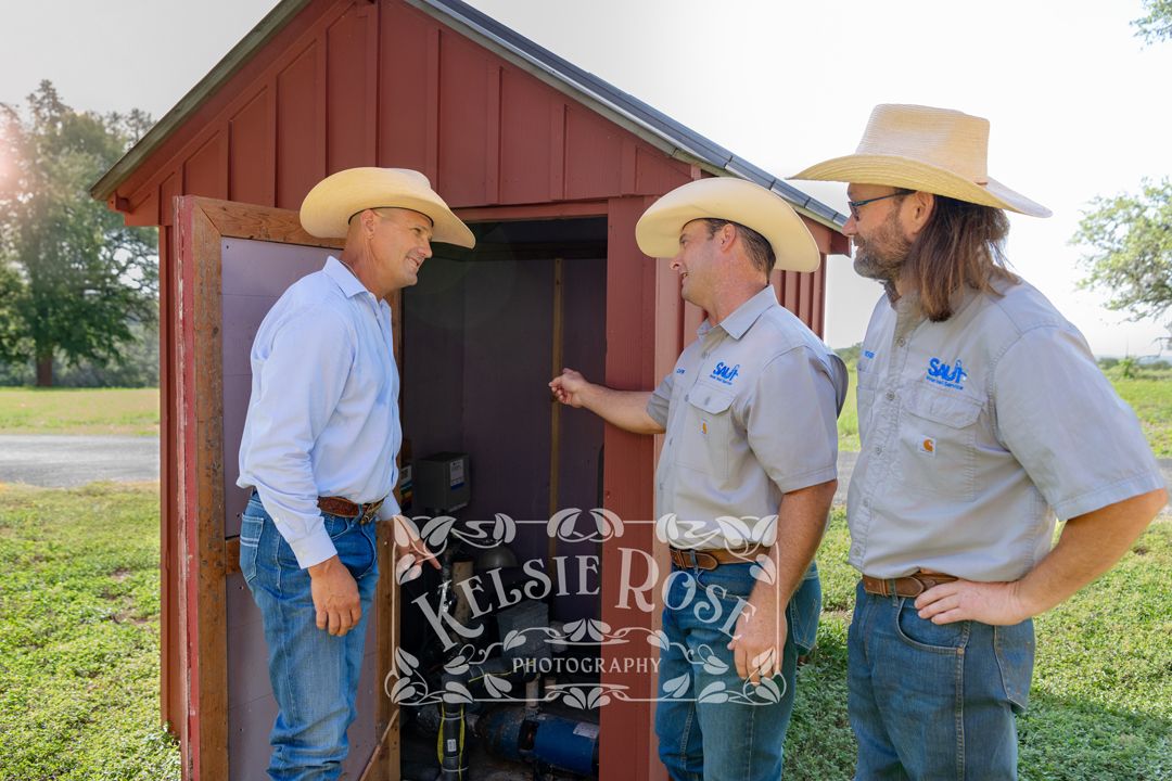 Three men in cowboy hats conversing near a red well building on a ranch in Texas.
