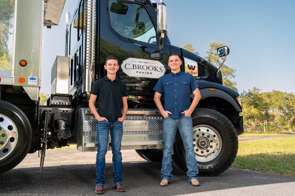 Father and son stand confidently in front of a C. Brooks Paving branded commercial truck outdoors.