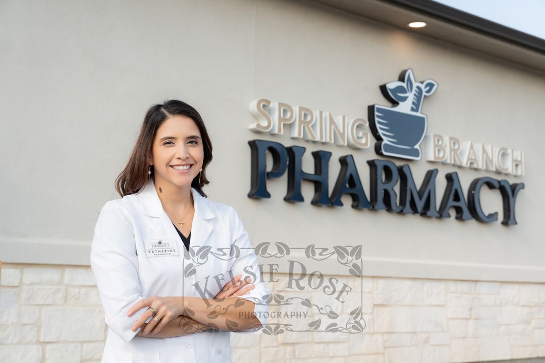 Female pharmacist in white lab coat standing confidently outside Spring Branch Pharmacy sign.