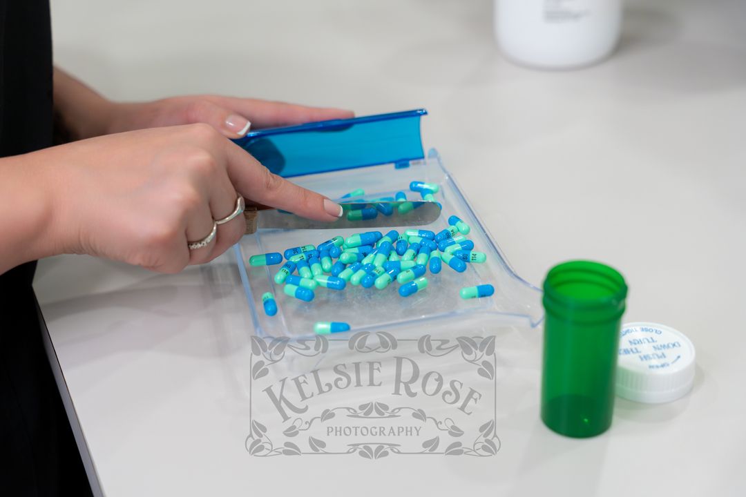 Close-up of hands sorting blue and aqua capsules into a pill organizer tray next to a green pill bottle on a white counter.