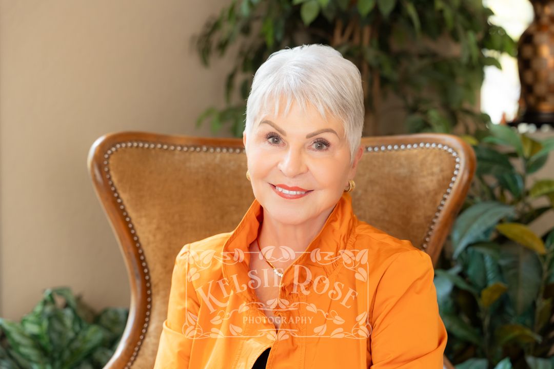 Elegant senior woman with short white hair wearing an orange blazer, seated in a wicker chair surrounded by green plants.
