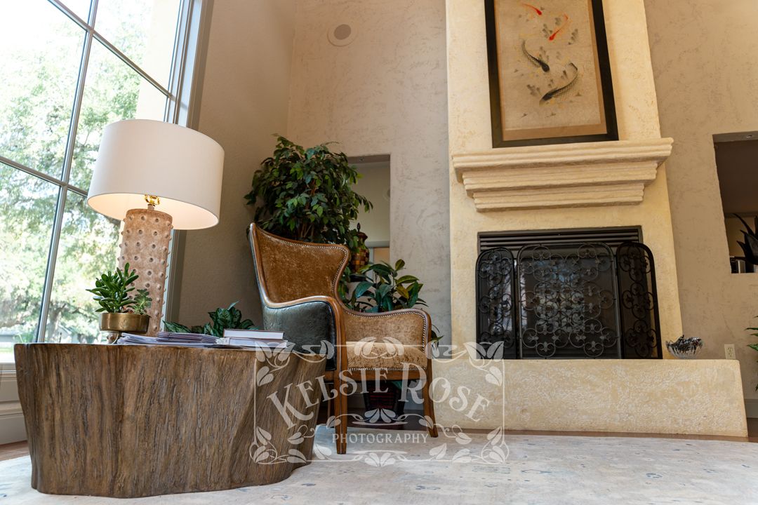 Elegant living room with a stone fireplace, wingback chair, wooden table, lamp, and lush greenery near tall windows.