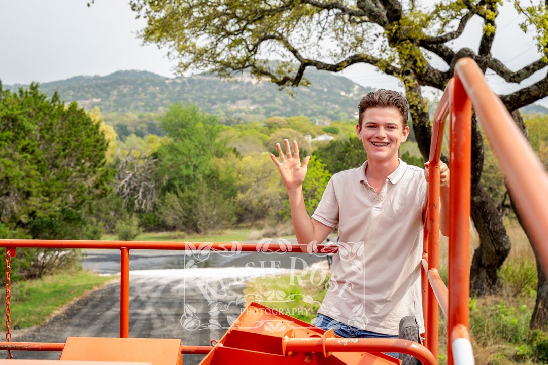 Young man waving from atop an orange paving machine with Texas hill country backdrop.