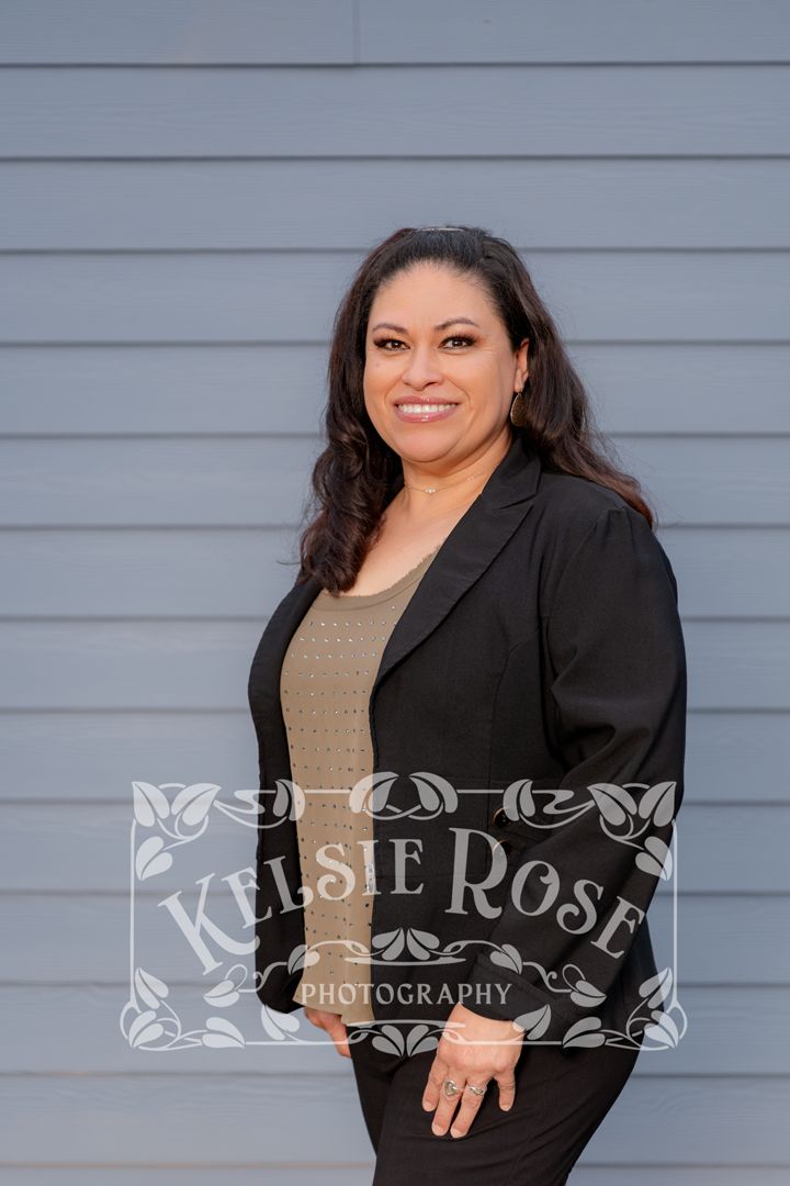 Professional portrait of businesswoman in black blazer and olive green top smiling against gray siding backdrop.