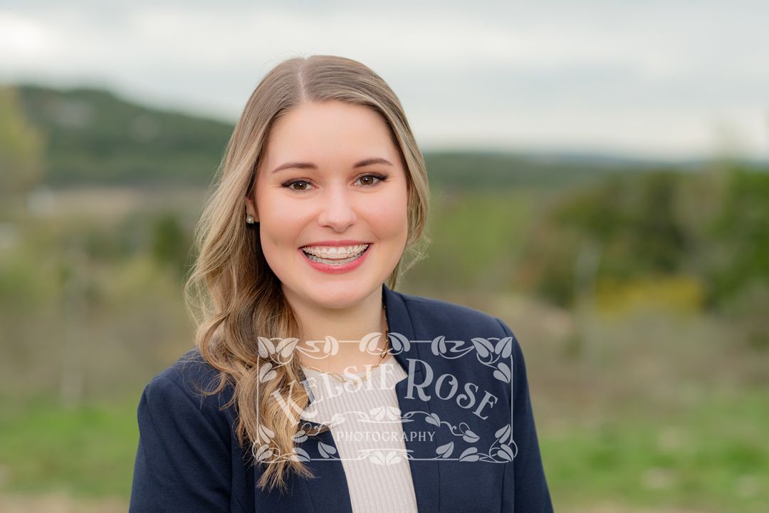 Headshot of young blonde woman in navy blazer smiling outdoors with Texas hill country background.