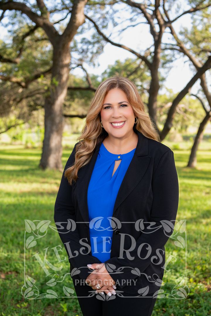 Professional portrait of smiling woman in blue blouse and black blazer posing outdoors under oak trees.