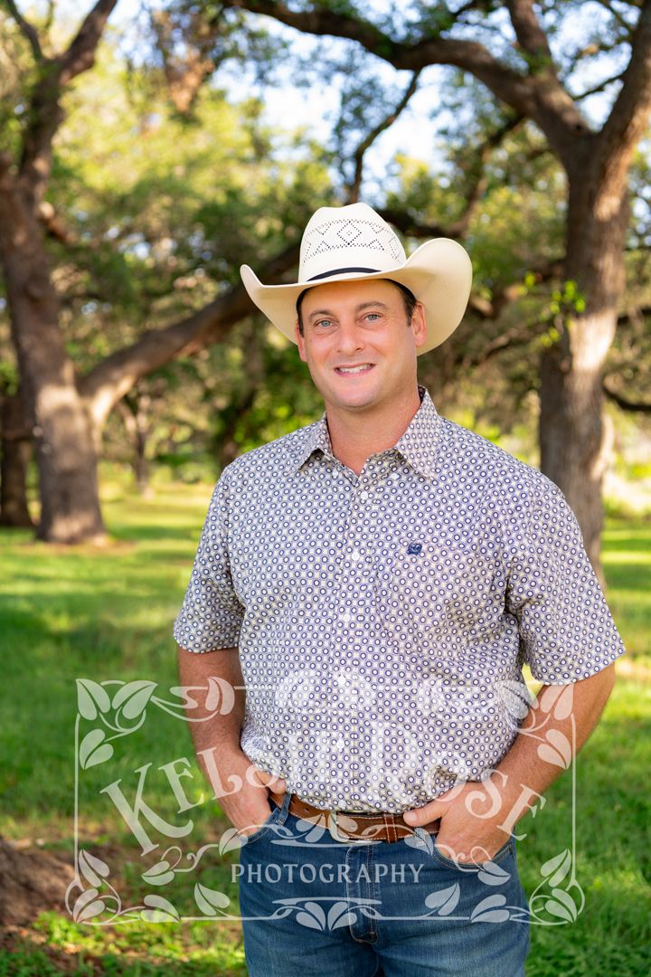 Professional portrait of smiling man in white cowboy hat and patterned shirt posing outdoors under oak trees.