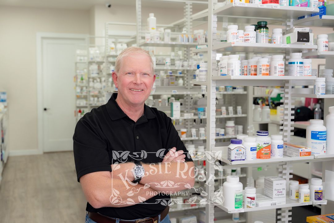 Smiling pharmacy employee in black polo shirt standing in front of shelves stocked with medications in a pharmacy.