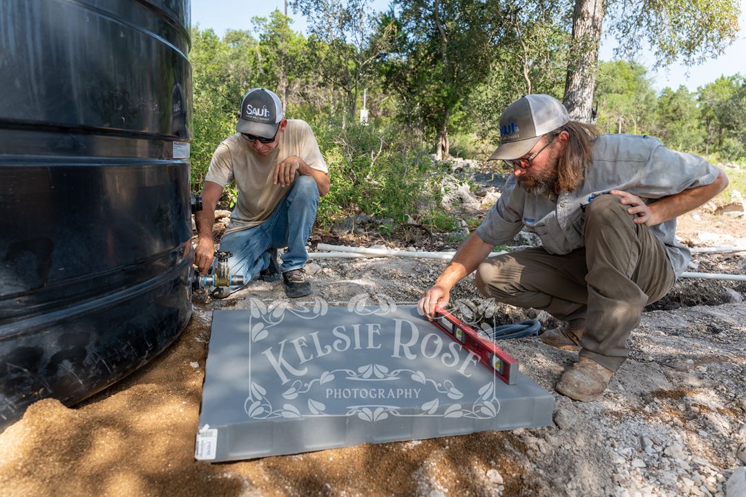 Two men installing a large black water storage tank and using a level on a gray platform on rocky ground outdoors.