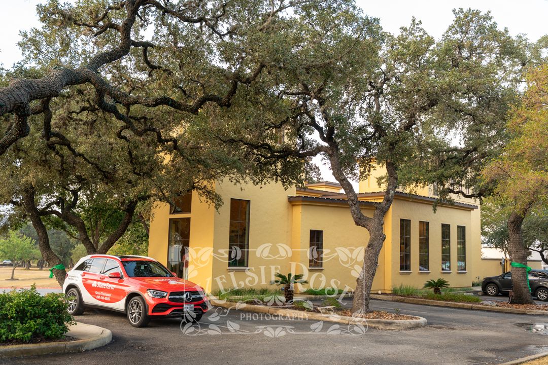 Yellow State Farm office building exterior with large oak trees and branded red Mercedes SUV parked outside.