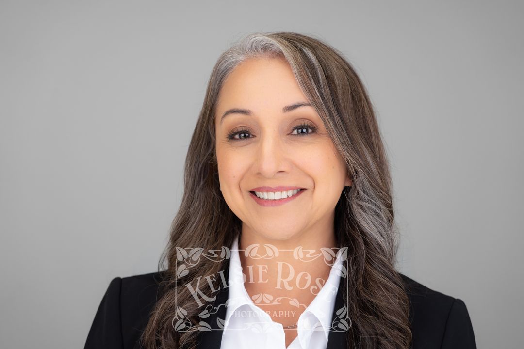 Professional headshot of a smiling middle-aged woman in a black blazer with gray-streaked hair against a gray background.