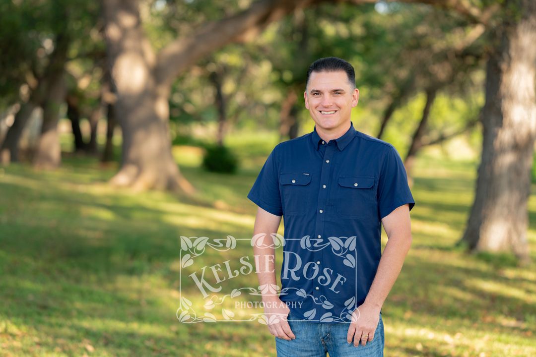 Portrait of smiling man in a navy blue shirt standing among green trees in a park.