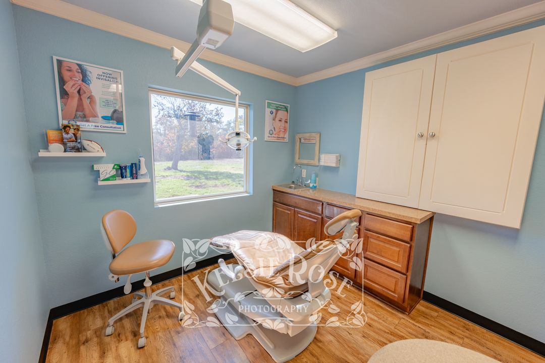 Light blue dental treatment room with patient chair, overhead light, wooden cabinets, and a window view of the Texas hill country.