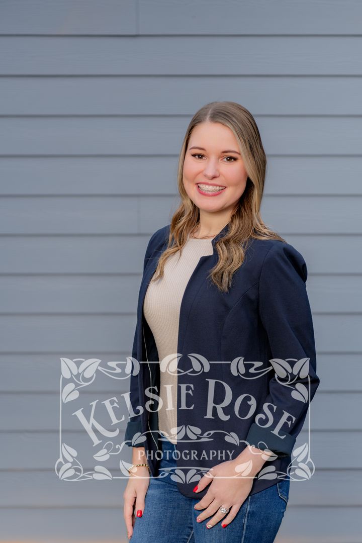 Young woman smiling in navy blazer and jeans against gray siding backdrop, professional headshot photography.