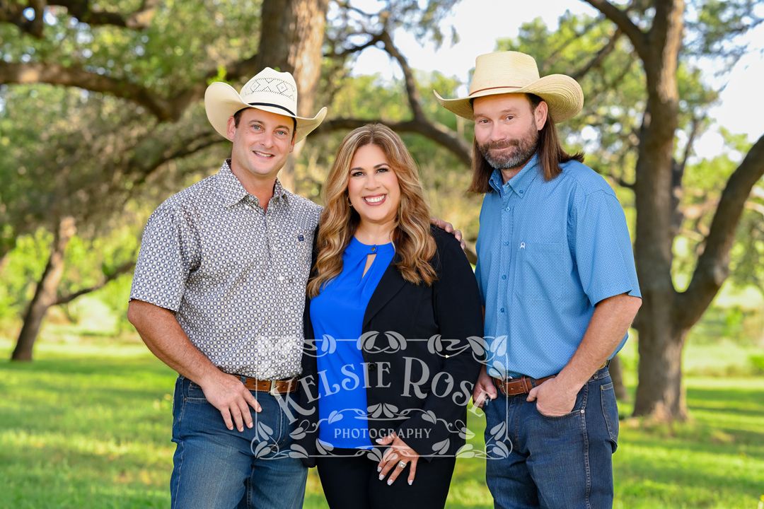 Three people posing outdoors, two men in cowboy hats flanking a woman in blue top and black blazer.