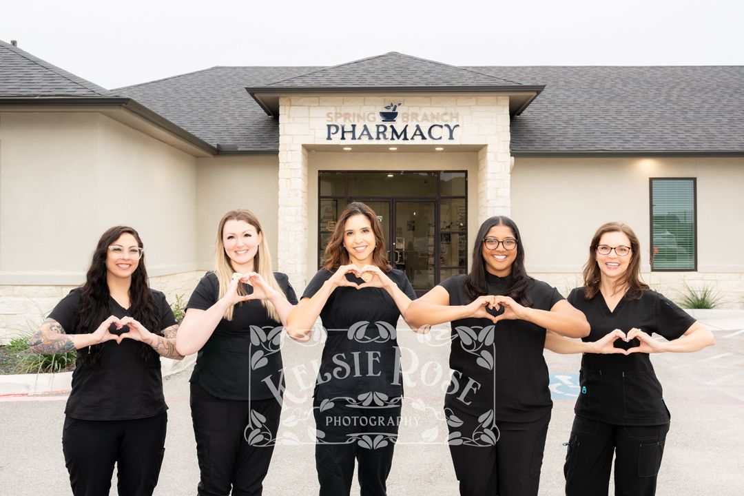 Five pharmacy staff members in black scrubs pose making heart shapes with their hands in front of a pharmacy.