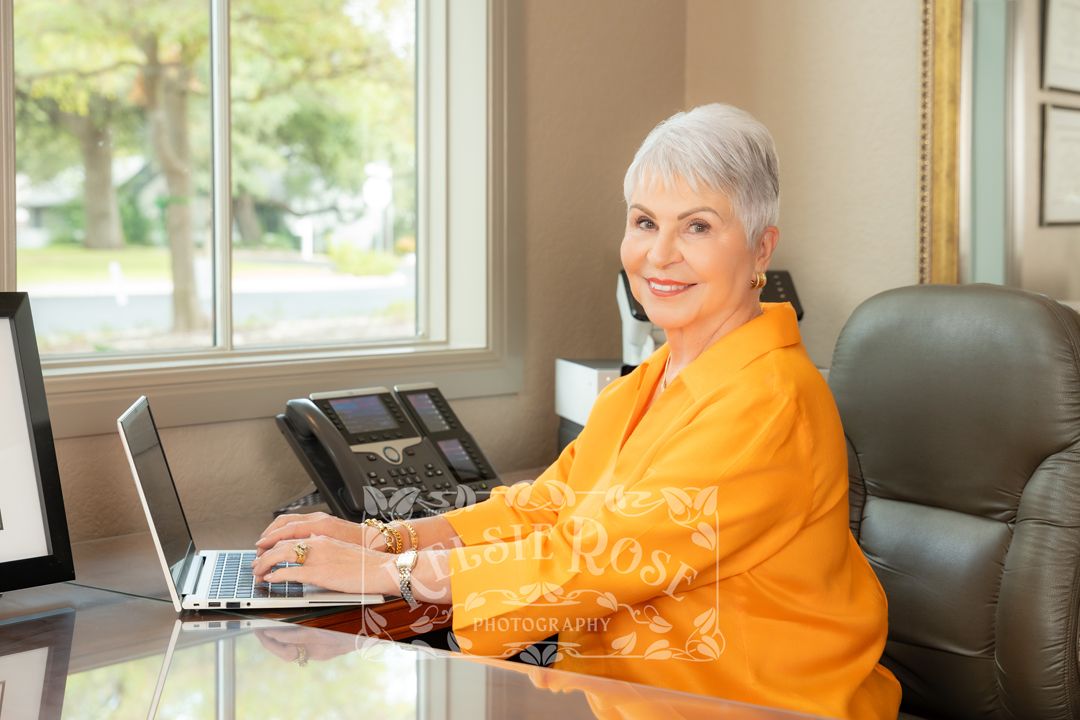 Smiling senior woman in yellow jacket working on laptop at desk with phone in bright home office.