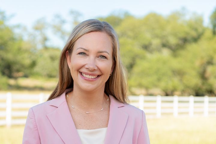 Headshot of smiling blonde woman in pink blazer poses outdoors near white fence with field in background.