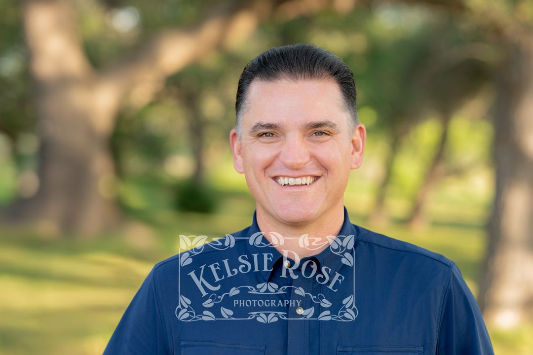 Headshot of a smiling man in a navy shirt with trees in the background.