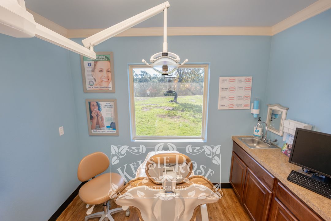 Bright dental treatment room with blue walls, patient chair, overhead light, computer, and garden window view.