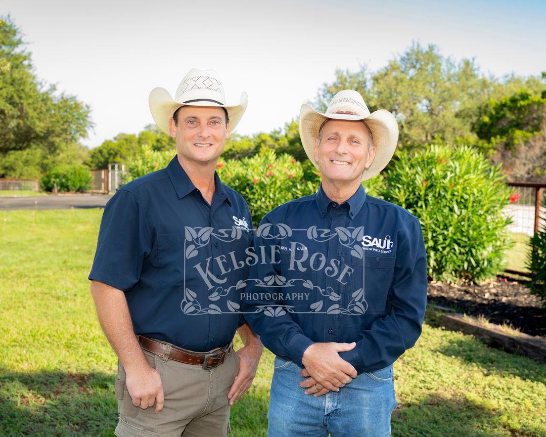 Two men in white cowboy hats and navy shirts smiling outdoors on a sunny day in a rural Texas setting.
