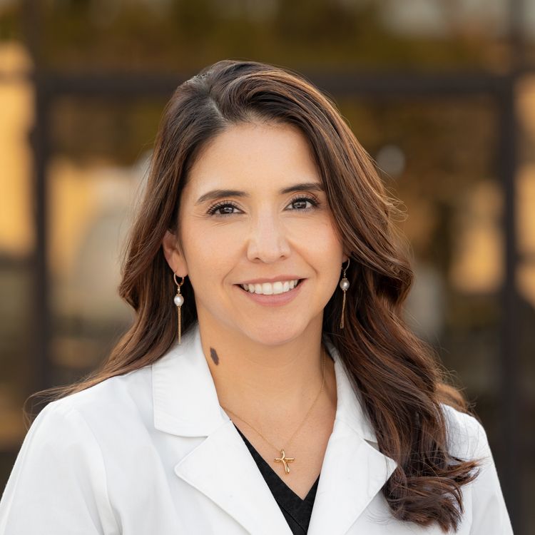 Professional woman in white lab coat smiling outdoors with brown hair and gold earrings.