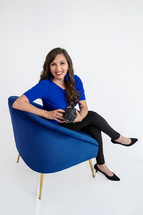 Businesswoman in royal blue top holding camera seated in blue velvet chair posing for professional branding photo.