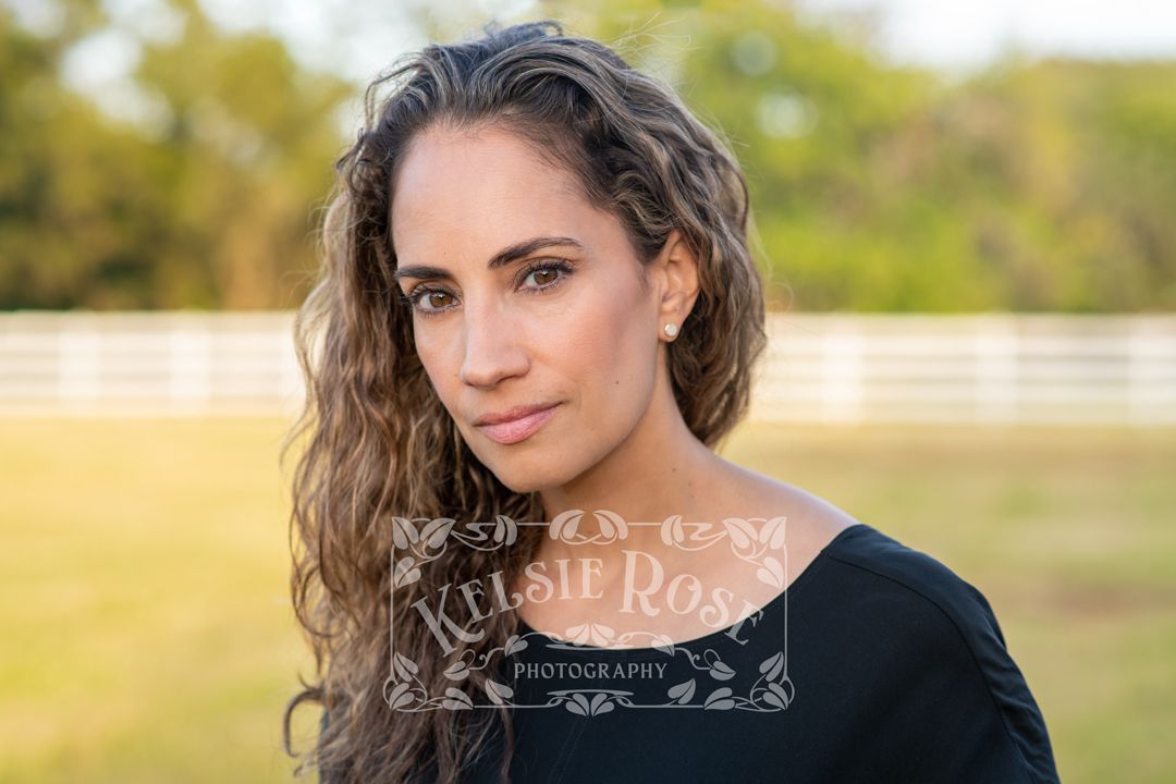 Headshot of a woman with long wavy hair in a black top, posing outdoors near a white fence.
