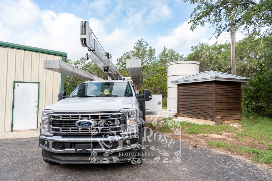 Water well service company truck with crane parked near metal building, water storage tank, and well building.