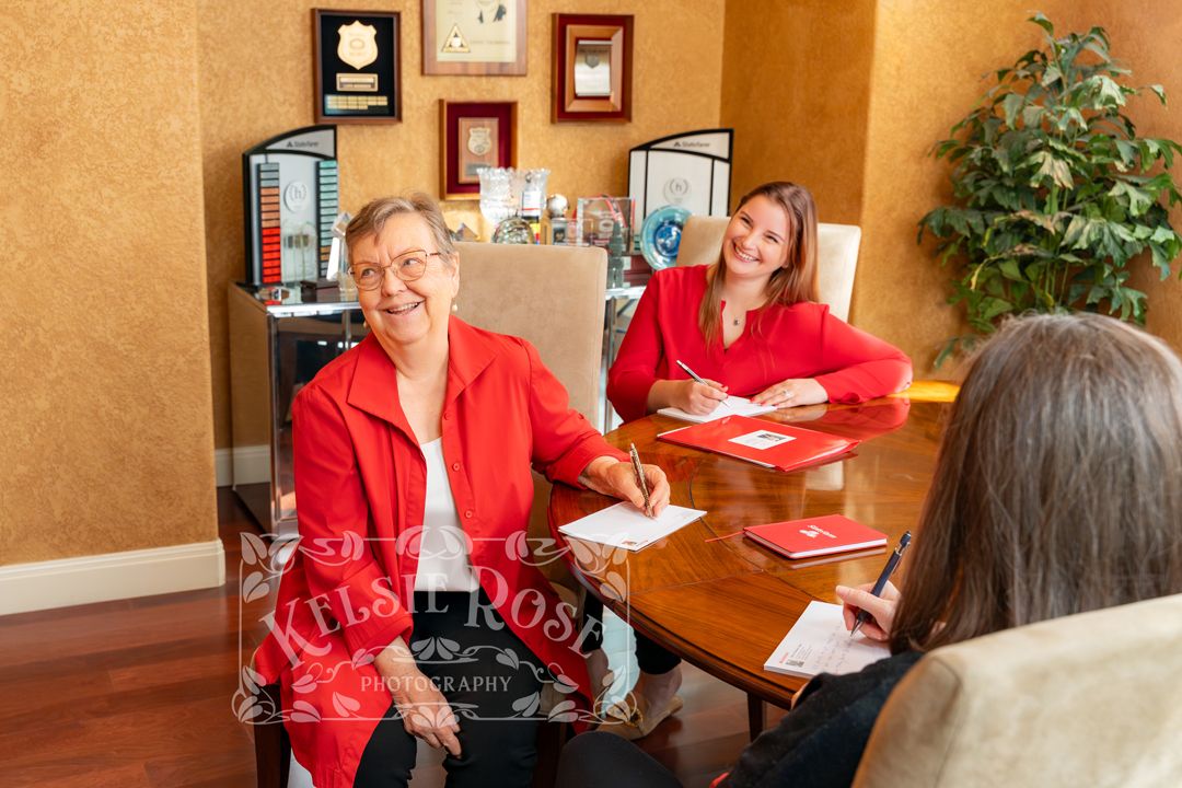 Two professional women in red discuss documents at a wooden conference table in a well-decorated office.