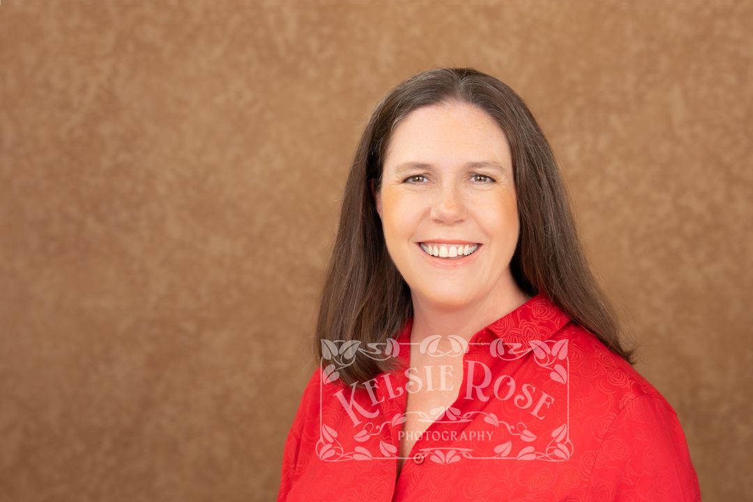 Woman with long brown hair smiling in red top against brown background, professional headshot photography.