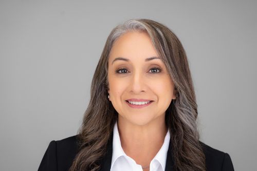 Professional headshot of businesswoman in black blazer smiling confidently, with long dark hair highlighted by natural gray streaks.